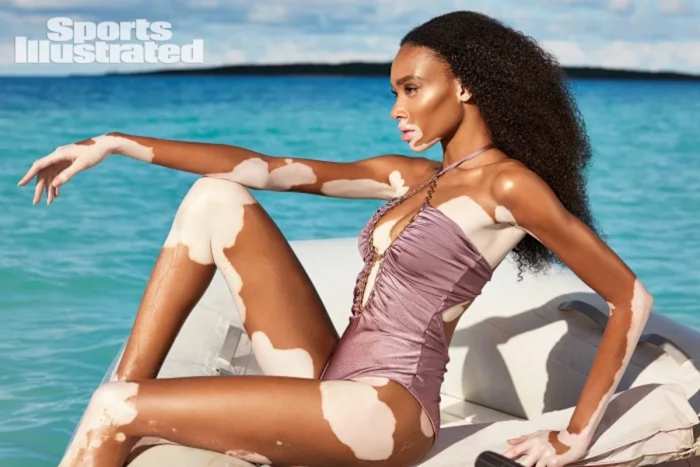 Winnie Harlow sits on a white raft wearing a lilac halter neck one-piece swimsuit.